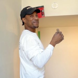 James Butler, owner of JB Paint, Tile and Landscaping LLC, turns back and smiles at the camera while painting a warm white wall