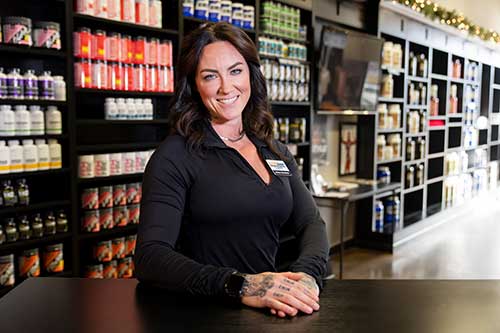 Local business owner, Amber Goodwin, stands smiling behind dark checkout desk at her Columbia, MO, business with supplement bottles in the background.