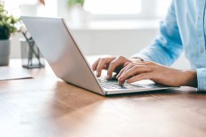 Man is typing while checking his email on silver laptop, atop a wooden desk with a white window in the background