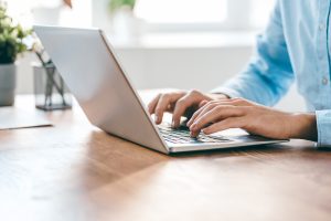banking fraud prevention in Columbia, MO -- a man types on a silver laptop on a light-colored wood desk with a white window in the background