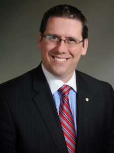 Brian Cox, Director of Training & Development - Brian sits smiling with a dark blue suit, red tie, white shirt, and light gray background