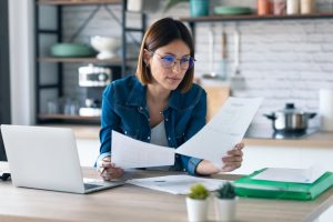 Business Banking in Ashland, MO | Shot of pretty young business woman working with computer while consulting some invoices and documents in the kitchen at home.